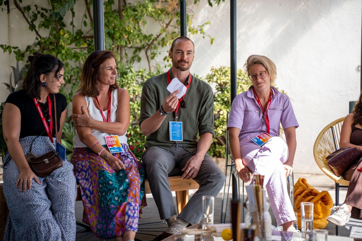 A participant holding up a folded paper as he speaks, while others on the bench listen, at Lisboa 2036